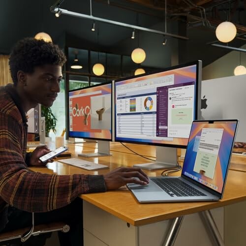 Person working at a desk with multiple computer screens and a laptop, showcasing the laptop's portability.